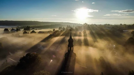 Luftaufnahme von Schönau-Berzdorf mit Nebel und Sonnenstrahlen, im Vordergrund ein Kirchturm.